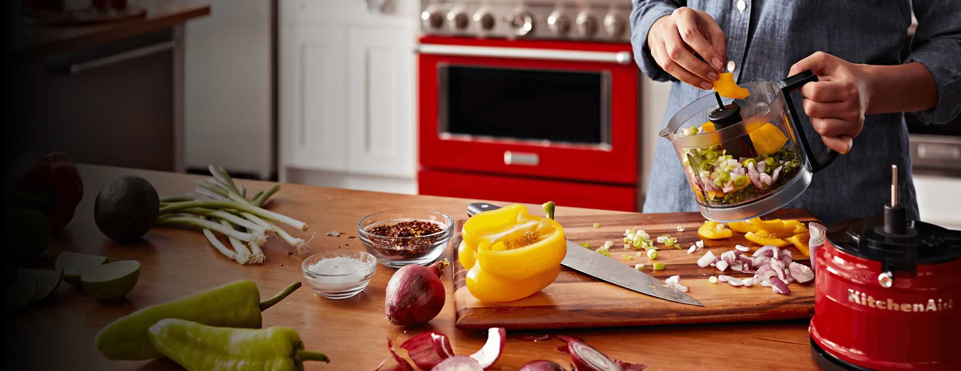 A KitchenAid food processor, loaded with a variety of colorful vegetables. All around it are the signs of preparation, with cut and uncut ingredients in various states of preparation.