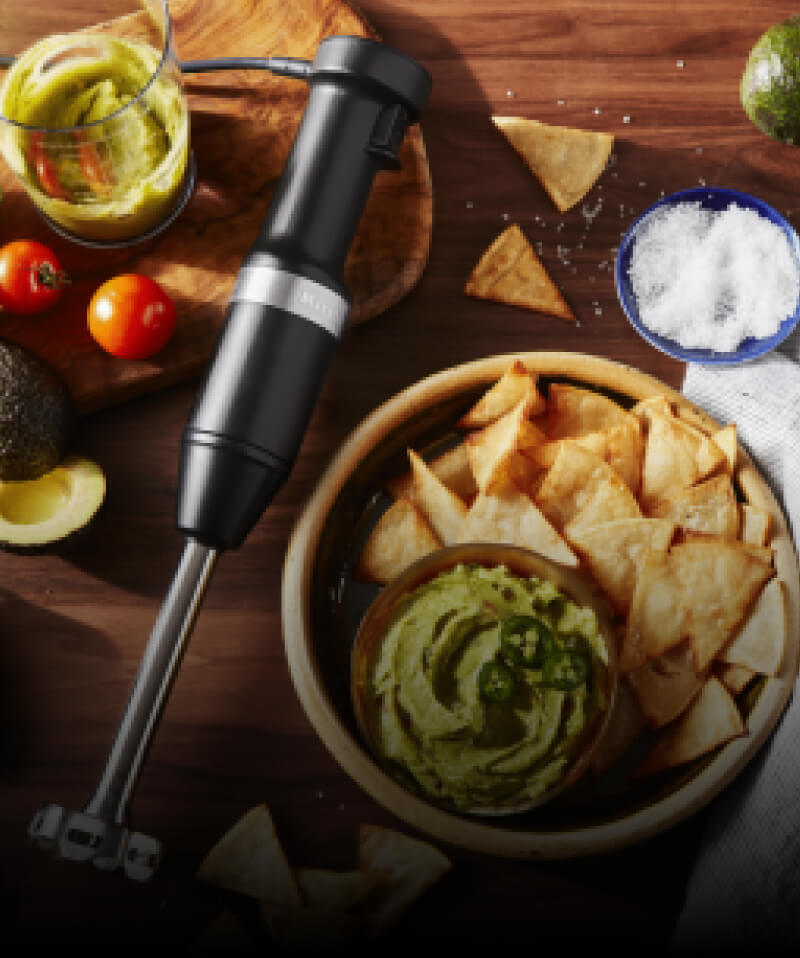 A KitchenAid® Hand Blender on a cutting board with different ingredients, bowls and chips. 