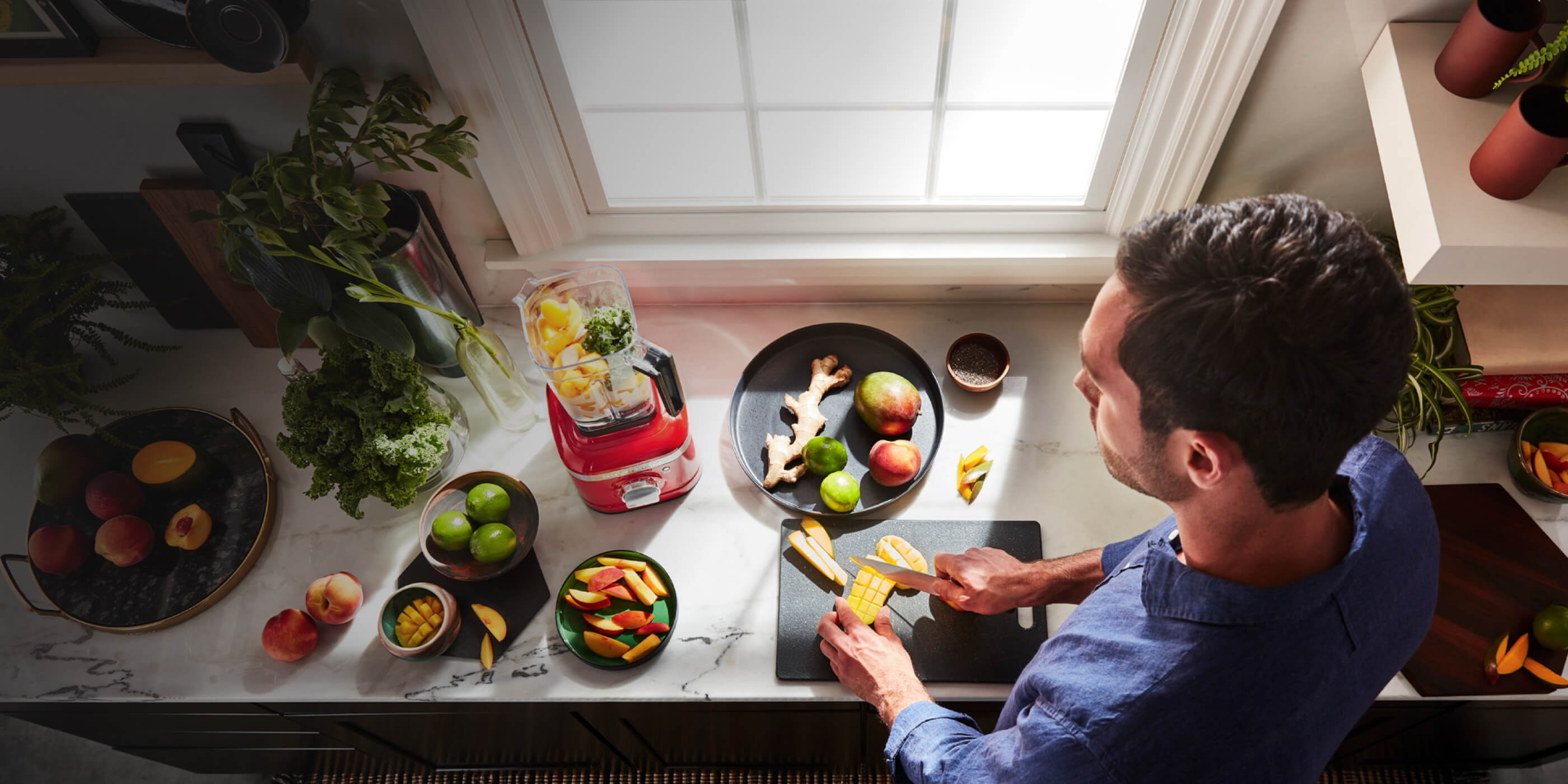 Overhead shot of a man cutting up a variety of fruits and vegetables to prepare a smoothie, some of which are already inside the blender.