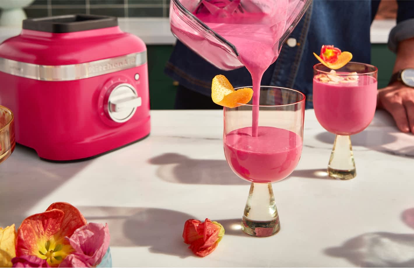 A person pouring a hibiscus blended drink into stylish, modern glasses with floral garnishes.