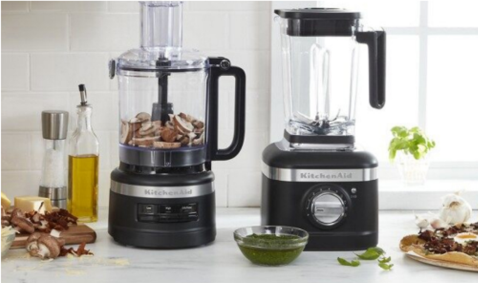 A KitchenAid food processor and blender on a countertop with different ingredients scattered around the counter.