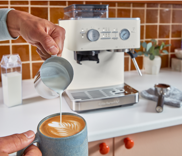 A person pouring steamed milk into their cappuccino, with a KitchenAid® Espresso machine in the background.