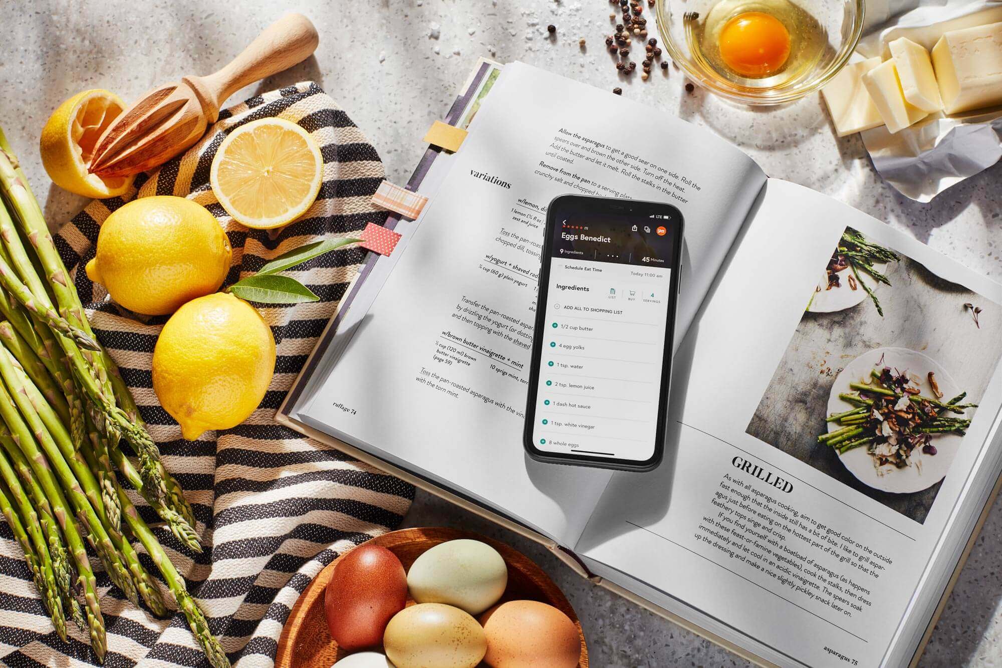 A phone opened up to an eggs benedict recipe resting on an open book's grilled asparagus recipe