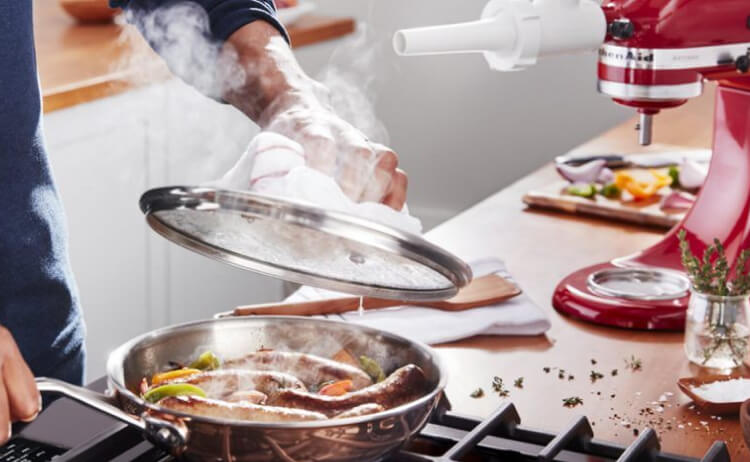 A man cooking juicy bratwursts with sautéed vegetables in a stainless steel pan over a stove.