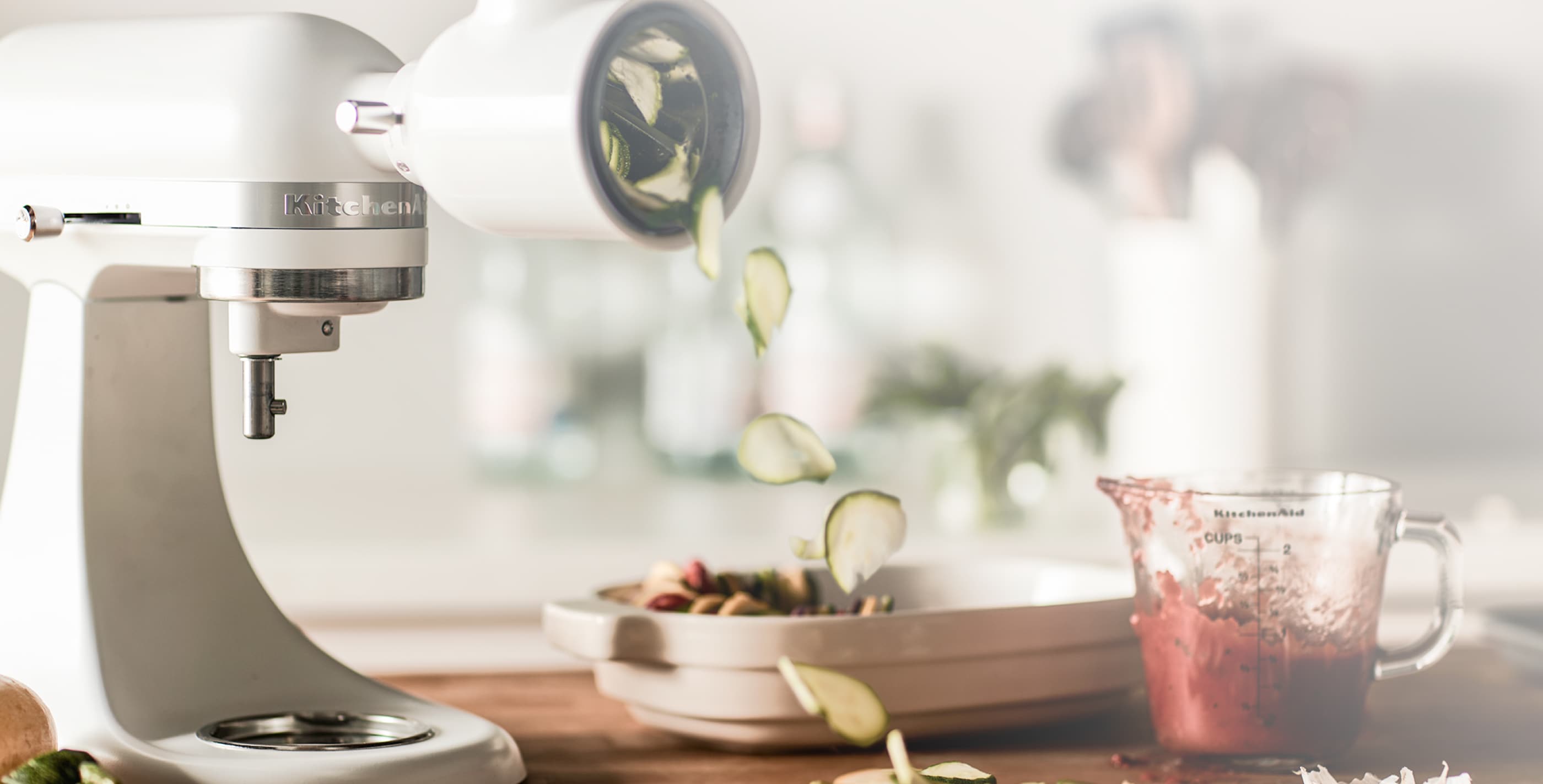 Image of cream-colored Stand Mixer with vegetable slicer attached, churning out cucumber slices.