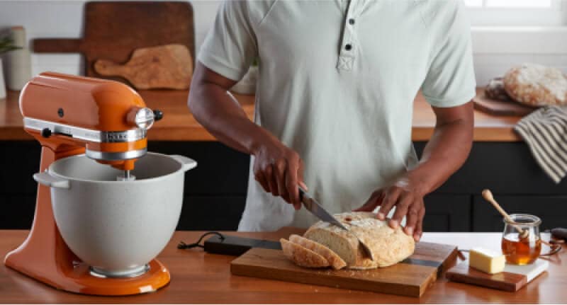 A person slicing a loaf of freshly baked bread, using a KitchenAid® Stand Mixer and the bread baking attachment.
