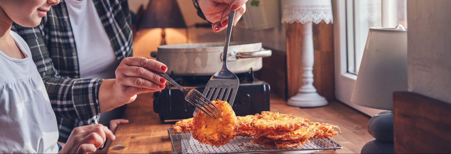 A young girl watching her mother gently move potato pancakes to a cooling rack.
