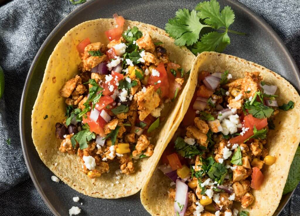 A plate of two chicken tacos topped with cilantro, tomato and onion garnished with queso fresco.