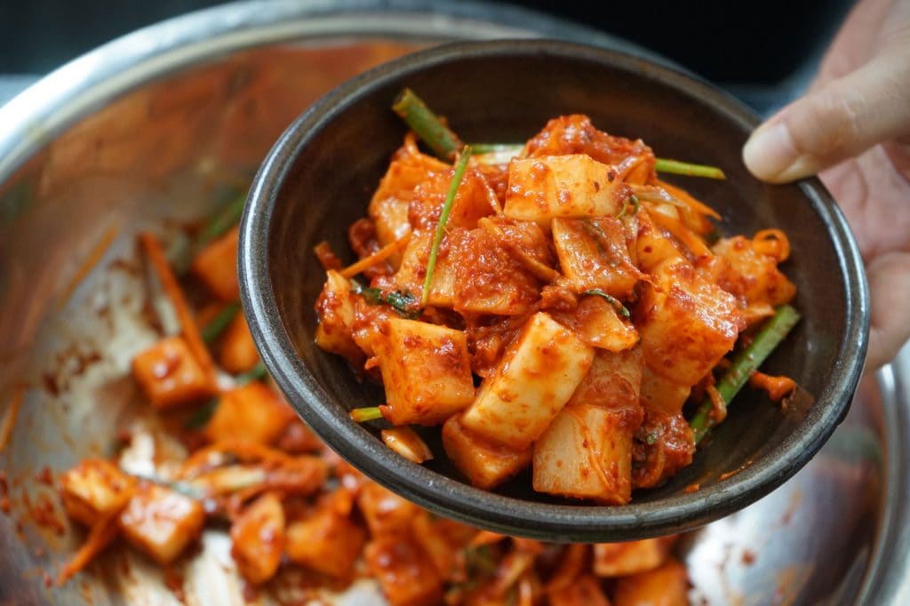 A person holding a small bowl filled with kimchi from a larger, stainless steel bowl.