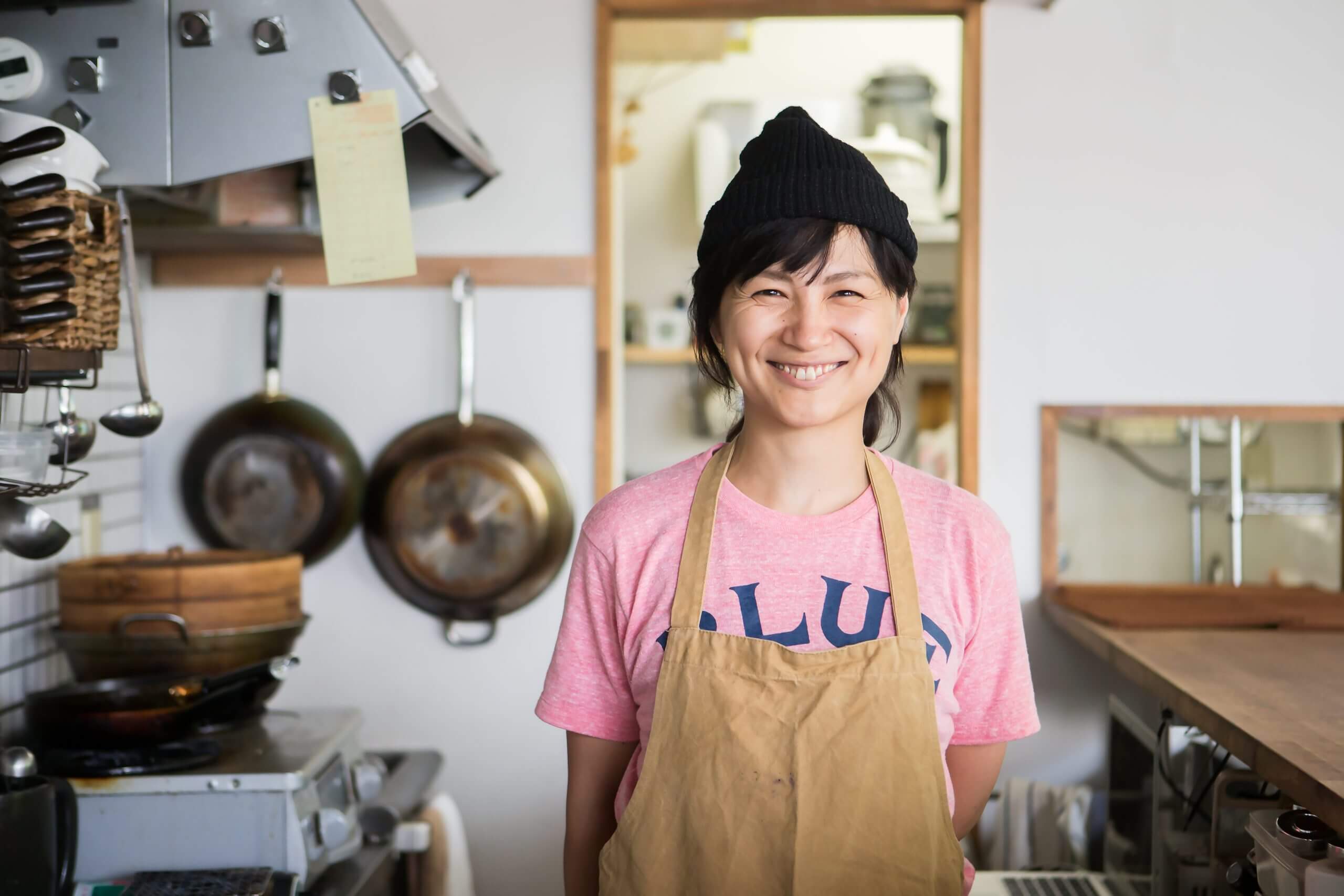 A woman in a canvas apron and pink shirt happily smiling at you from her kitchen.