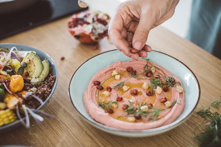 A soft hand sprinkling pomegranate on smooth hummus with herbs and whole chickpeas.