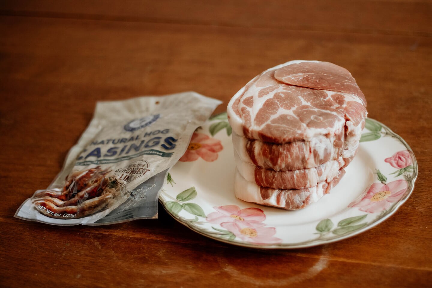 Meat resting on a floral china plate next to an unopened bag of natural hog casings.