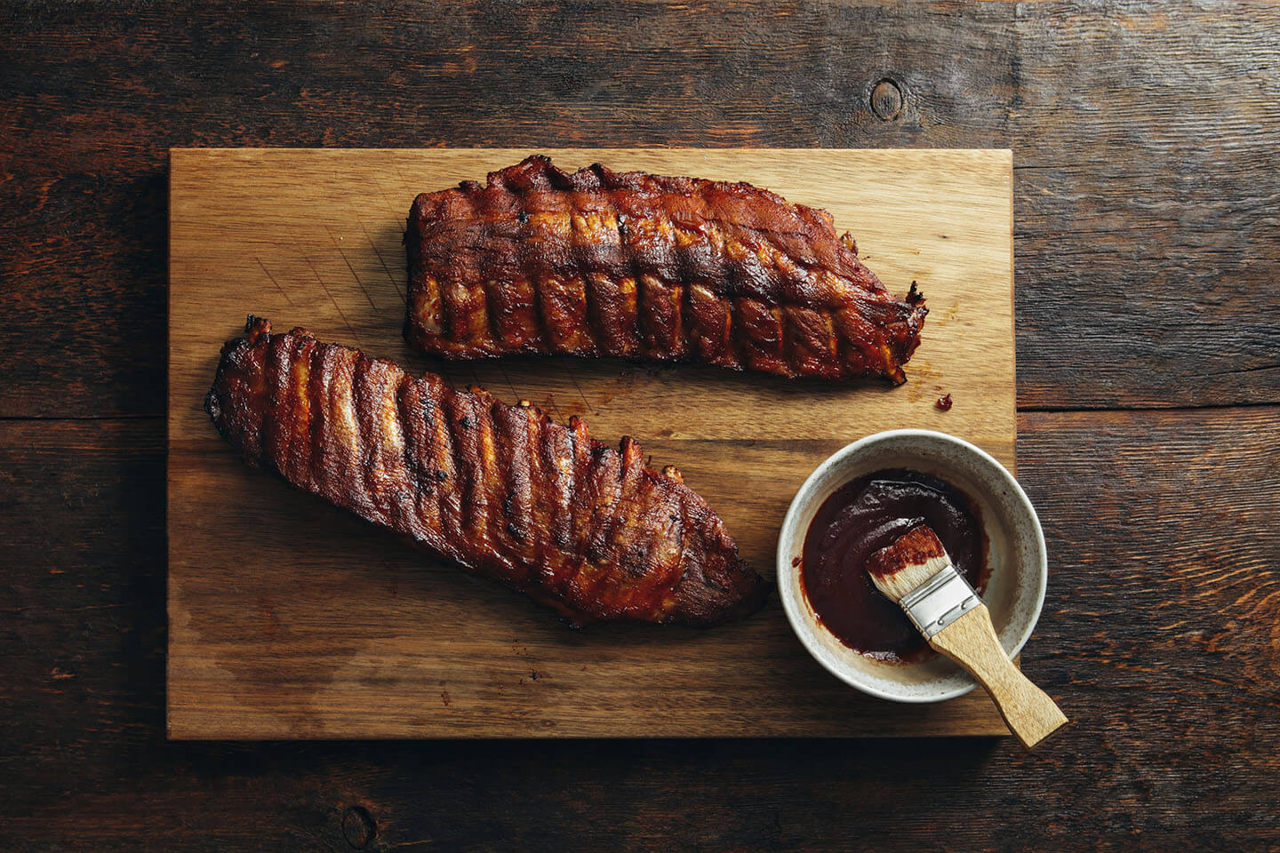 A rack of ribs resting on a wooden cutting board next to a bowl of barbeque sauce.