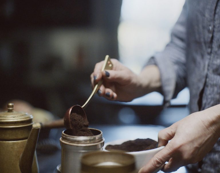 Erica pouring spoonfuls of ground coffee into a small metal carafe.