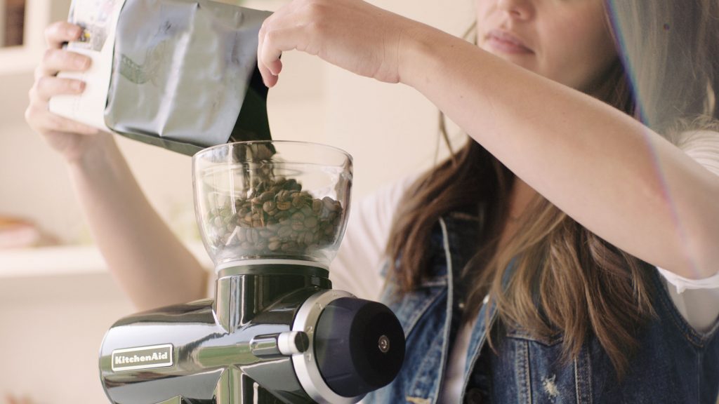 A young woman pouring a small bag of coffee beans into a KitchenAid® Burr Grinder.