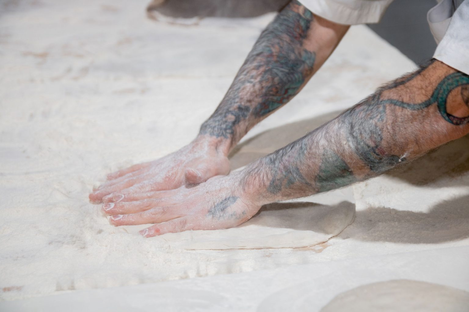 A person flattening dough surrounded by flour.