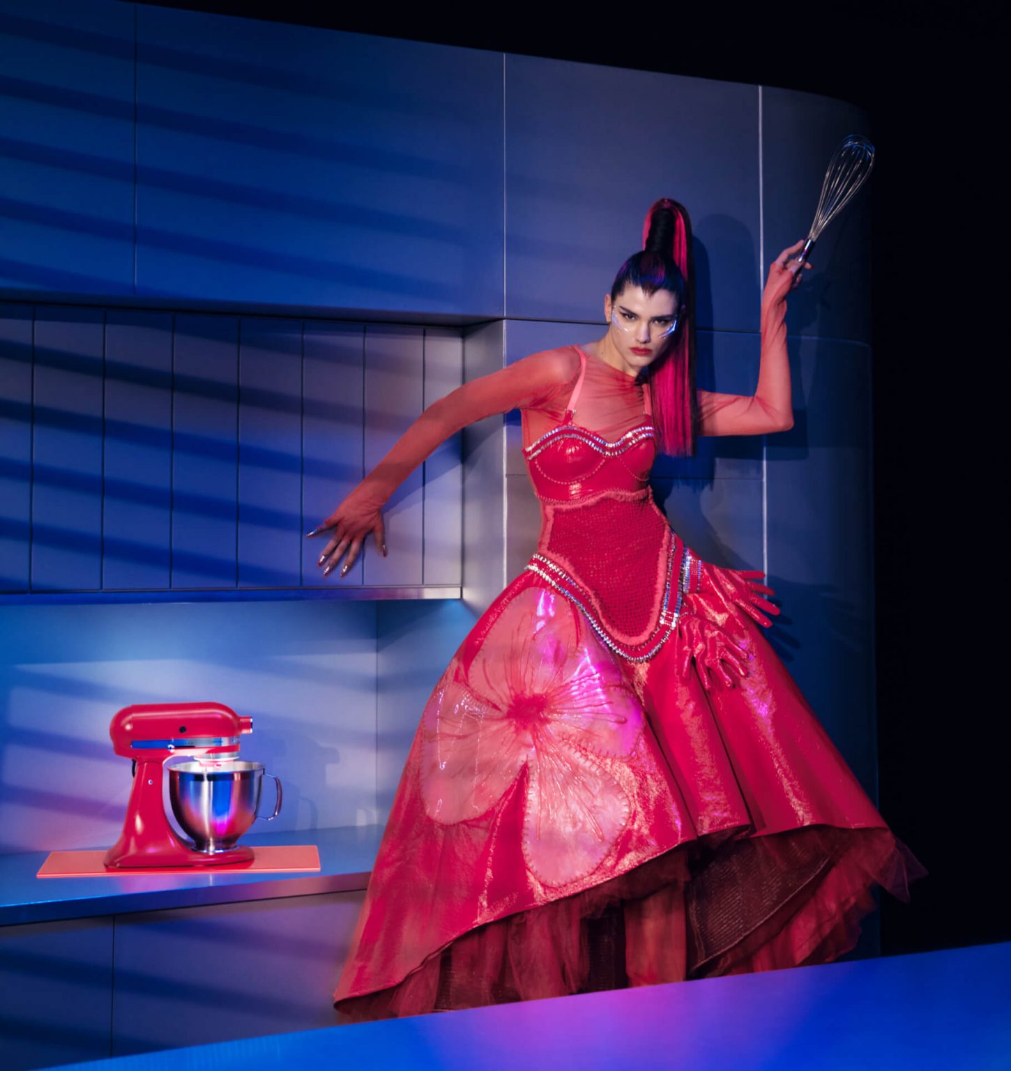 A model posing in a kitchen wearing Look Nine while holding a whisk next to the Artisan® Series Stand Mixer in Hibiscus on the countertop. 