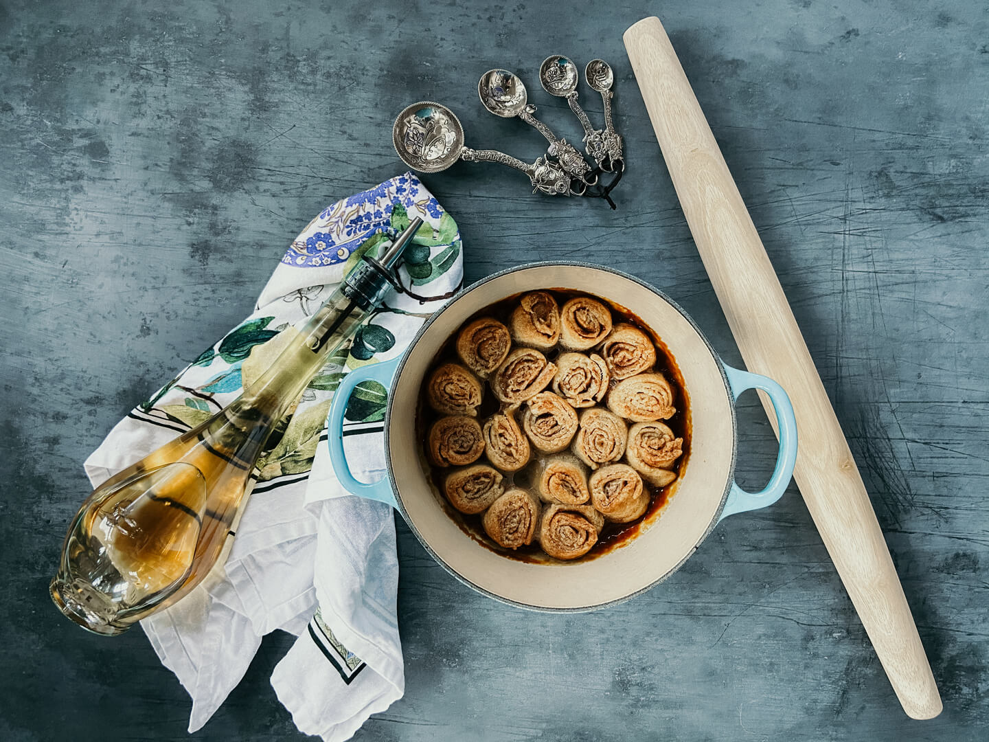 A light blue dutch oven holding cinnamon rolls next to a bottle of vinegar on a delicate towel.