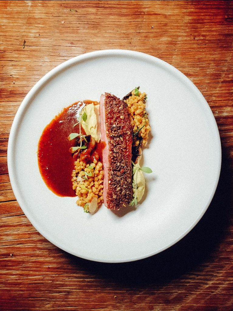 A slice of seared duck resting a bed of cous cous with sauce.