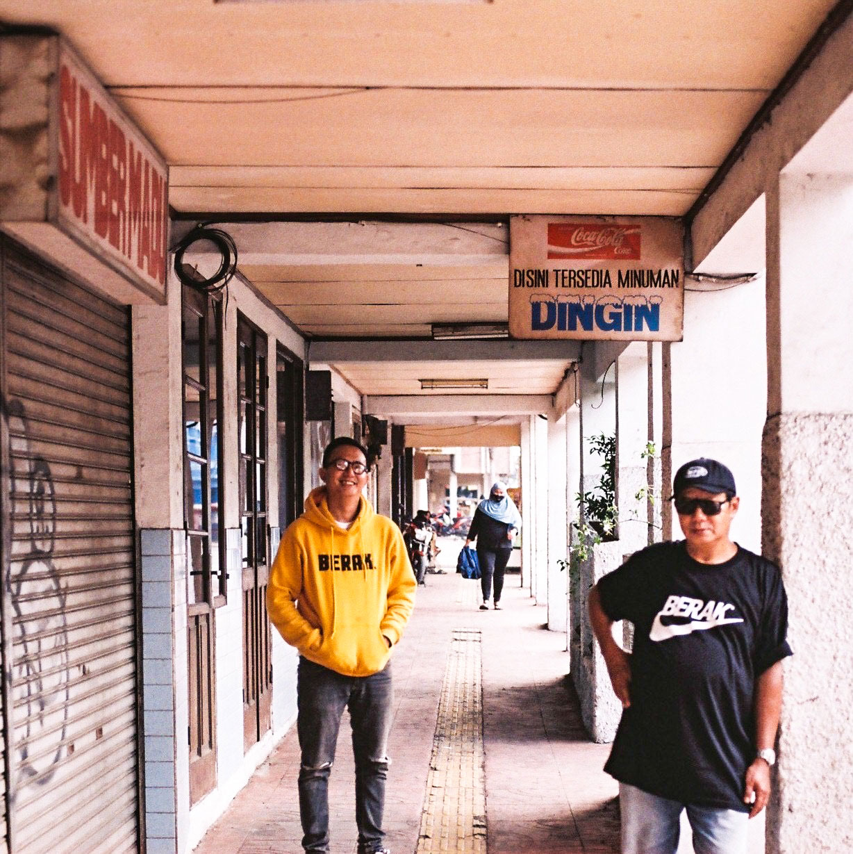 Paulus Supomo in black sunglasses leaning against a cement wall with son Yulius Iskandar in yellow sweatshirt standing behind.