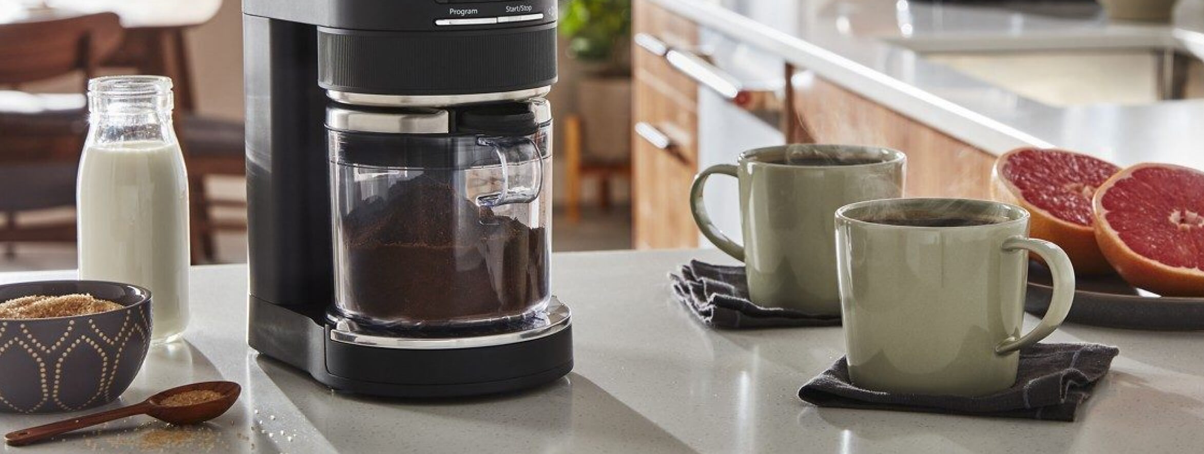 Best home coffee setup on countertop, including KitchenAid® Burr Grinder surrounded by milk, cinnamon, two cups of coffee and grapefruit.