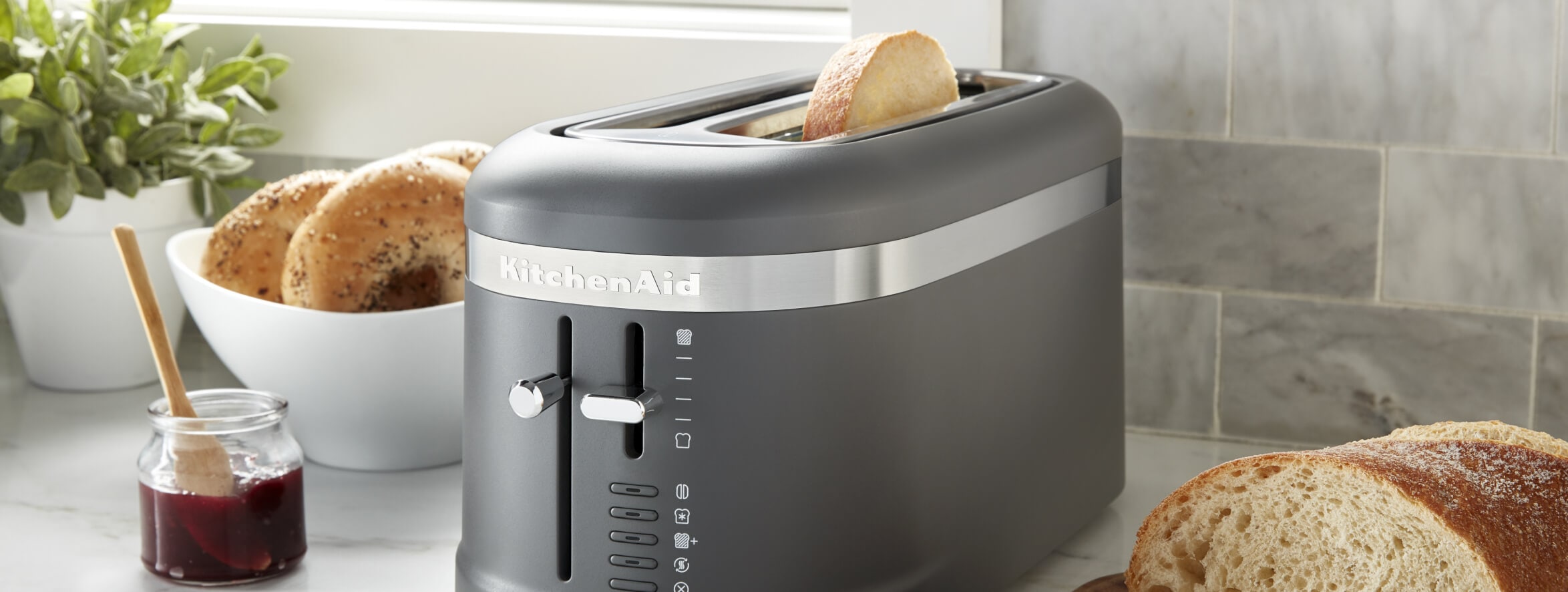 A matte black toaster on a counter with bagels, bread and jam