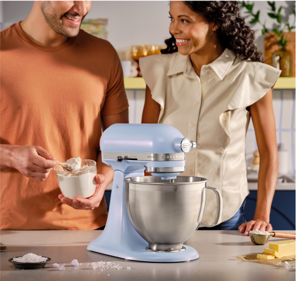 Two people smiling while adding dry ingredients to a Blue Salt stand mixer.