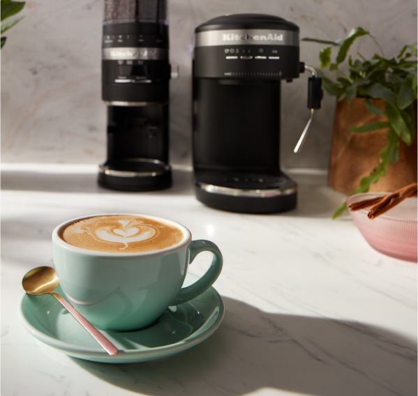 A cup of espresso in a light green mug in the foreground and KitchenAid® Espresso and Burr Coffee Grinder in the background.