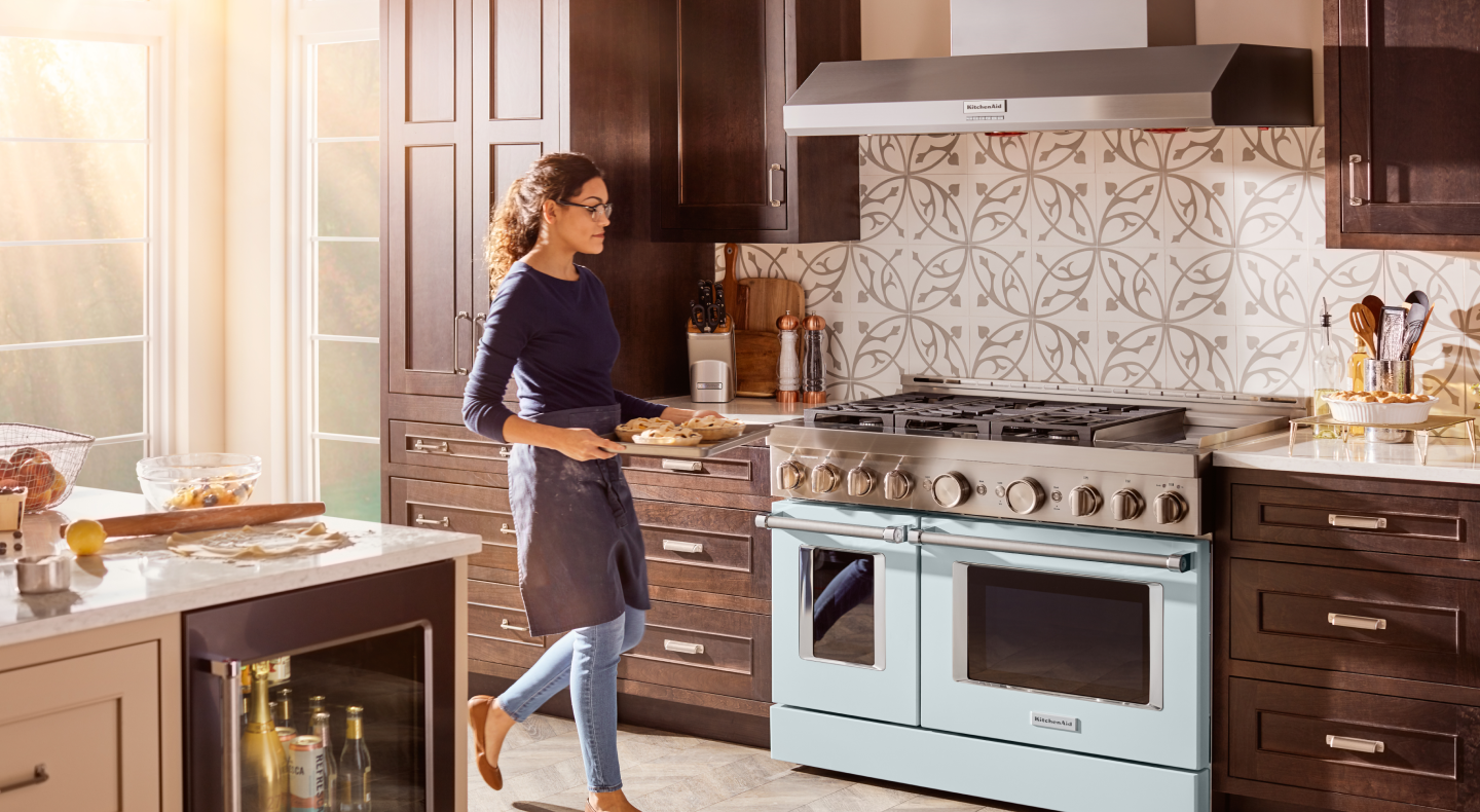 Person carrying tray of food toward a blue KitchenAid® commercial-style gas range Person carrying tray of food toward a blue KitchenAid® commercial-style gas range