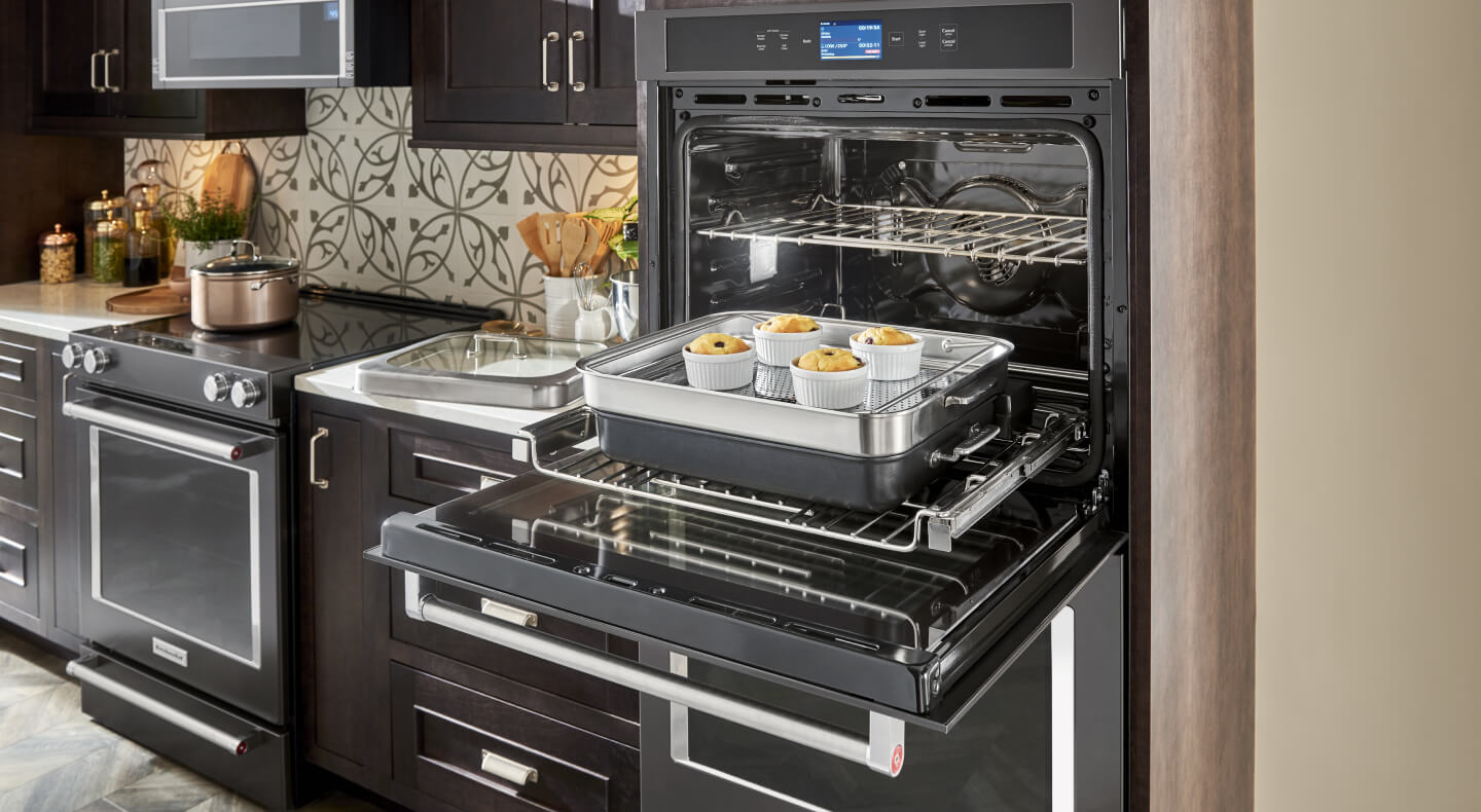A sleek, modern kitchen with dark appliances and a KitchenAid® Double Wall Oven, open, with the racks pulled out and desserts on a pan sitting on one rack