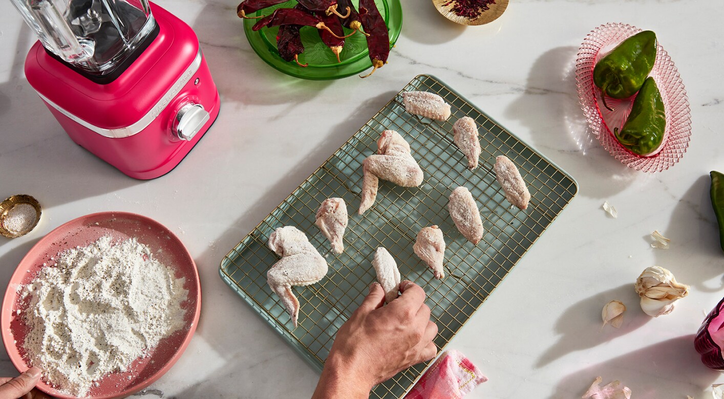 A person placing dredged and breaded chicken wings onto a wire rack over a baking sheet with different ingredients across the countertop. 