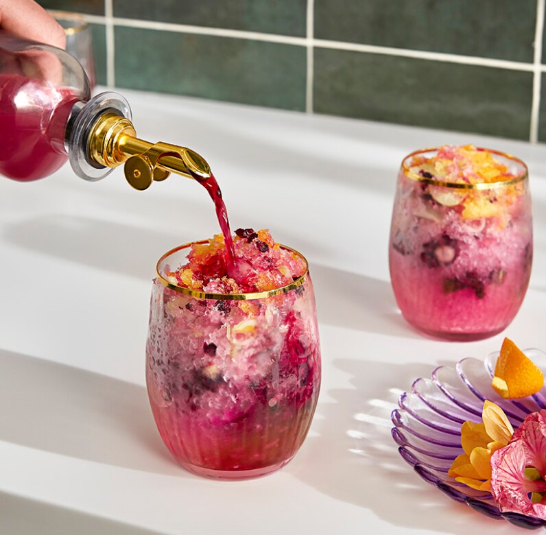 A person pouring hibiscus syrup into a pink shaved ice drink with floral garnishes.