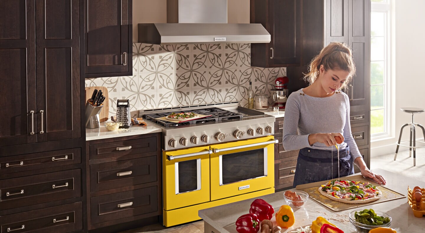 A woman prepping food at a countertop with a KitchenAid® 48” Commercial-Style Range in Yellow Pepper behind her