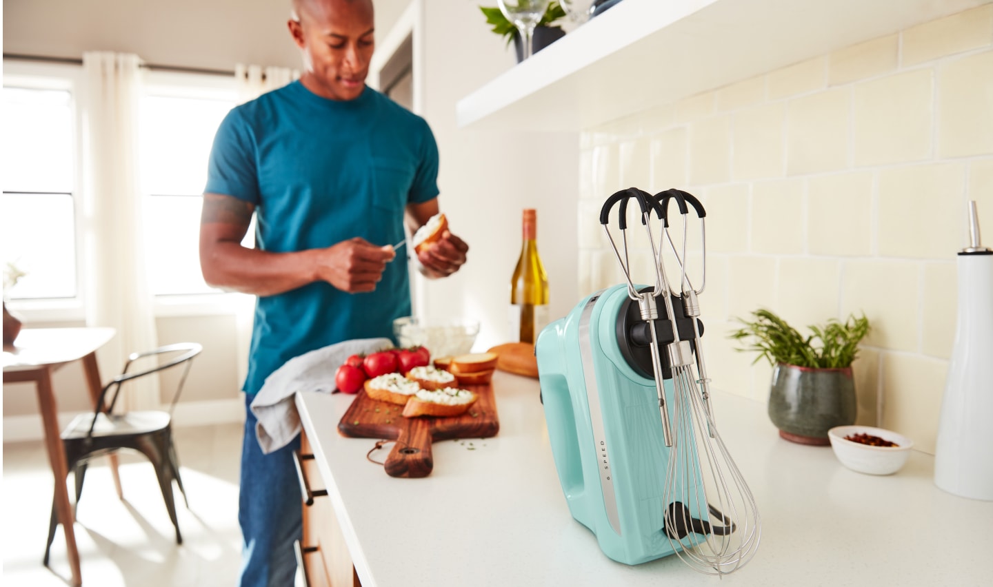 A man in a modern kitchen spreading goat cheese on a crostini next to a KitchenAid® hand mixer. A man in a modern kitchen spreading goat cheese on a crostini next to a KitchenAid® hand mixer.