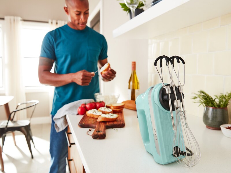 A man in a modern kitchen spreading goat cheese on a crostini next to a KitchenAid® hand mixer. A man in a modern kitchen spreading goat cheese on a crostini next to a KitchenAid® hand mixer.