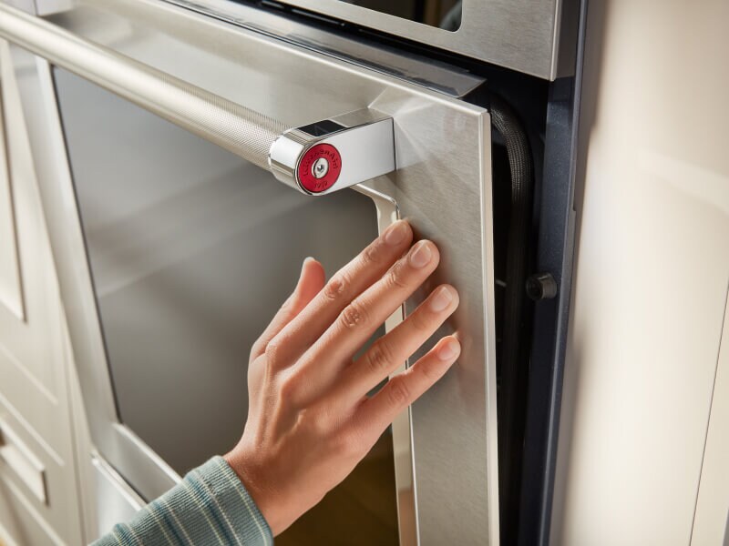 A hand touching a stainless steel oven door with a glass window in it