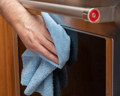 A close up image of oven door glass with a hand wiping it with a rag in the foreground