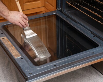 The inside of oven door glass with a hand painting a cleaning paste on it in the foreground