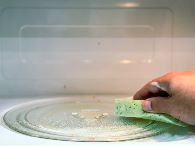 A hand using a sponge to clean the inside of a microwave oven