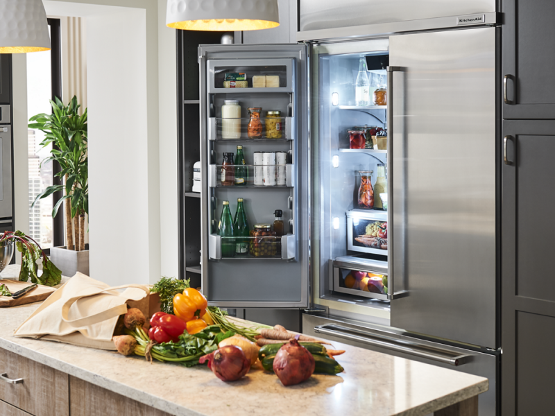 Fresh vegetables on countertop and a french door refrigerator behind with doors open