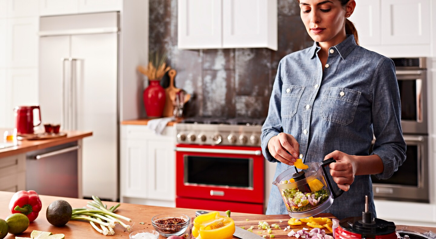 A woman standing at a modern kitchen counter putting vegetables in a KitchenAid® food chopper work bowl.  A woman standing at a modern kitchen counter putting vegetables in a KitchenAid® food chopper work bowl.