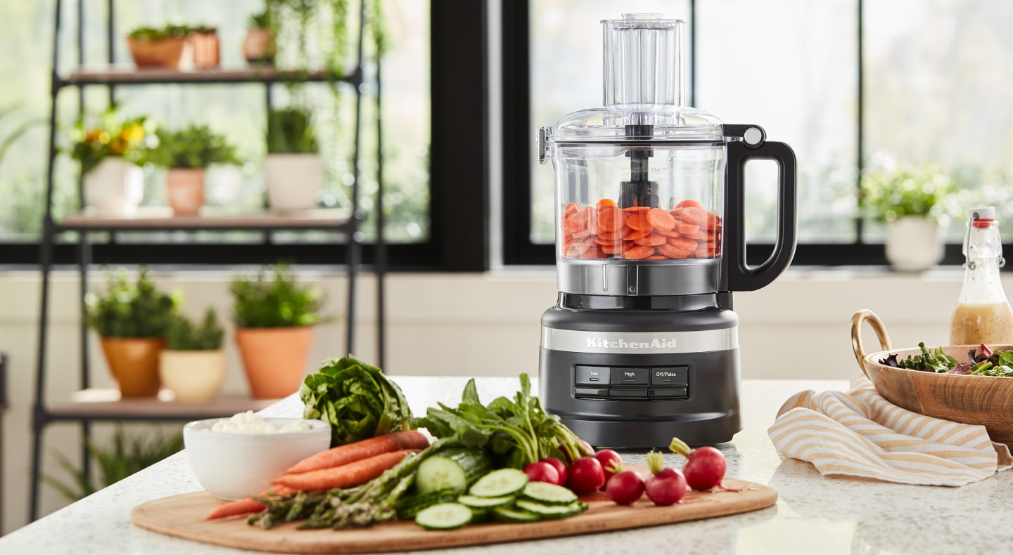 A KitchenAid® food processor mixing vegetables in a bright, modern kitchen. A KitchenAid® food processor mixing vegetables in a bright, modern kitchen.