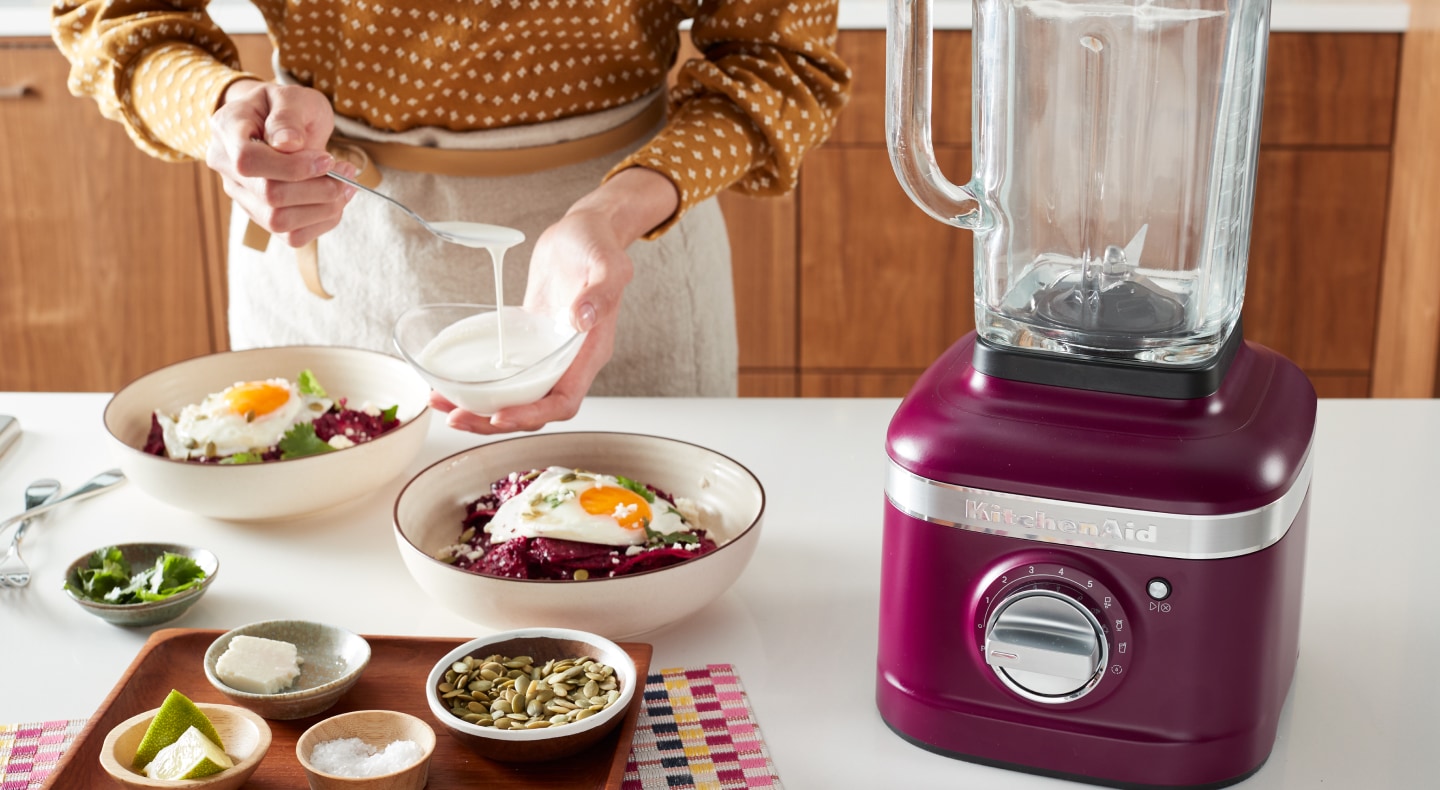 Person topping a harvest bowl with sauce next to a KitchenAid® blender Person topping a harvest bowl with sauce next to a KitchenAid® blender