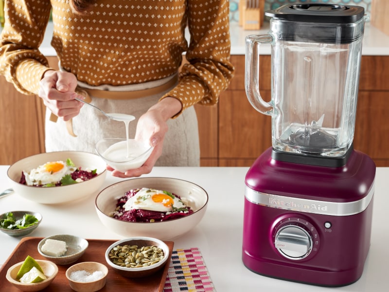 Person topping a harvest bowl with sauce next to a KitchenAid® blender Person topping a harvest bowl with sauce next to a KitchenAid® blender