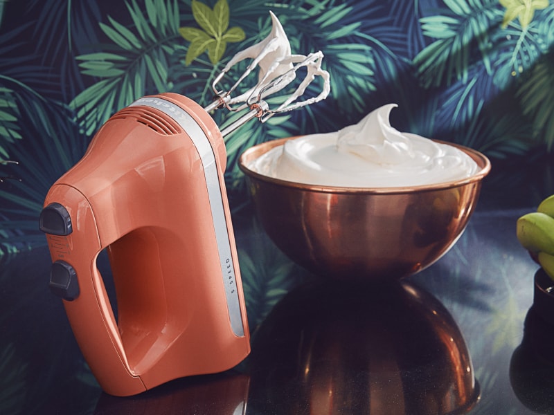 Orange KitchenAid® hand mixer next to a bowl of whipped cream Orange KitchenAid® hand mixer next to a bowl of whipped cream