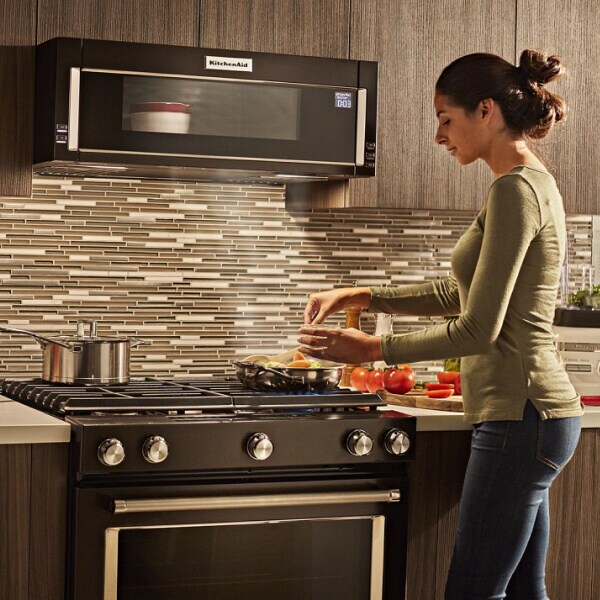 Person cooking food on a gas stovetop below a KitchenAid® low profile microwave warming food