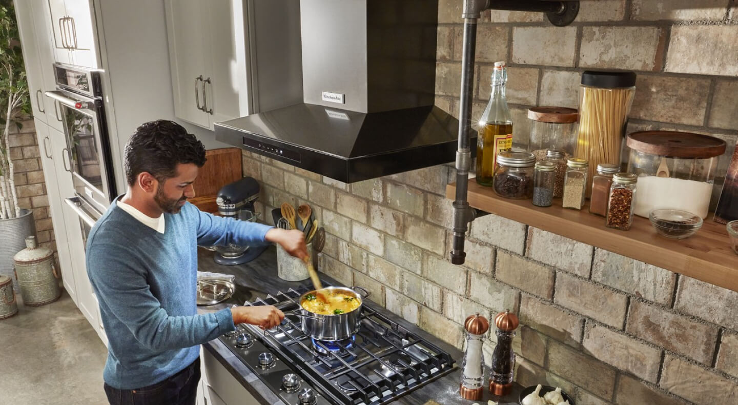 Person stirring food in pot below a KitchenAid® wall-mount canopy hood