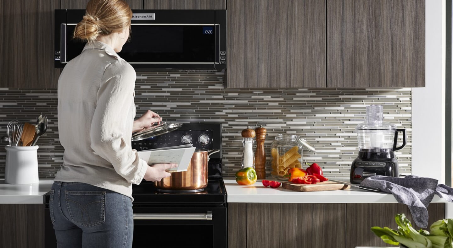 Person putting a lid on a stock pot beneath a black, KitchenAid® low profile microwave