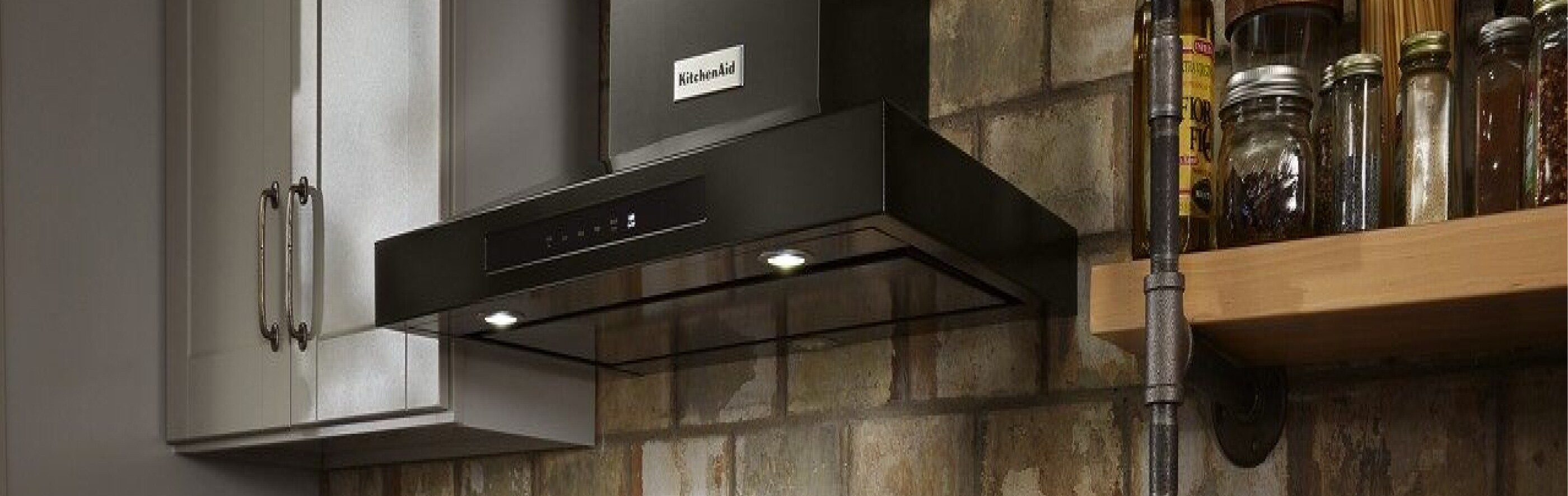 Black KitchenAid® wall mount hood in a kitchen in front of a brick wall