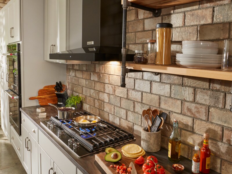 Black KitchenAid® canopy range hood over gas stovetop with brick backsplash Black KitchenAid® canopy range hood over gas stovetop with brick backsplash